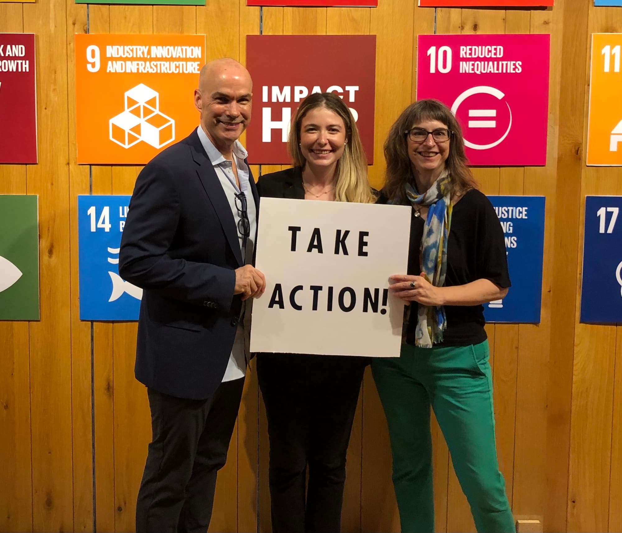 World Tree leadership holding a "Take Action!" sign in front of a wall of UN Sustainable Development Goal posters