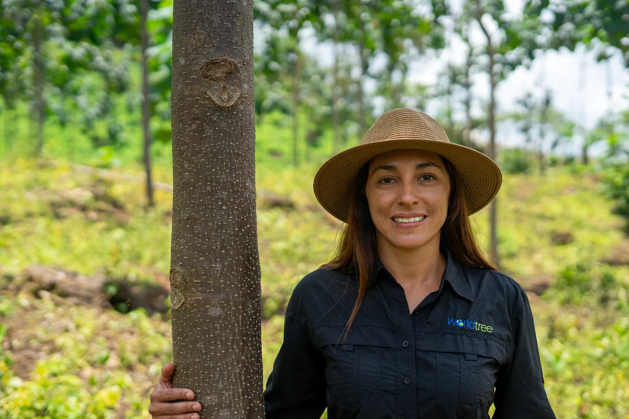 A World Tree team member in a branded shirt, smiling beside a young paulownia on a partner farm